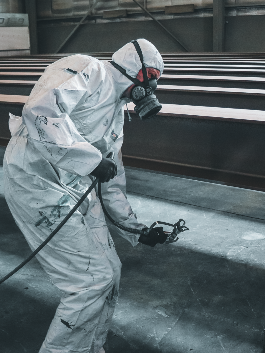 Worker in protective gear spray painting metal beams inside a factory setting.